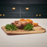 A crispy roast rests on the Cole & Mason Berden Acacia Carving Board, garnished with rosemary and Yorkshire puddings. Juice grooves catch drippings, complementing a kitchen with dark drawers and brass handles.