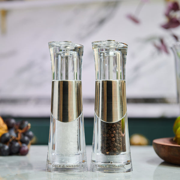 The Cole & Mason Bobbi Salt & Pepper Mill Set features two clear grinders with silver accents and a durable mechanism, displayed side by side—one filled with coarse salt, the other with black peppercorns against a softly blurred background.