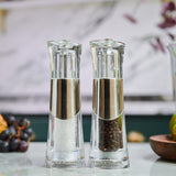 The Cole & Mason Bobbi Salt & Pepper Mill Set features two clear grinders with silver accents and a durable mechanism, displayed side by side—one filled with coarse salt, the other with black peppercorns against a softly blurred background.