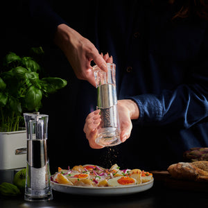 A person uses the Cole & Mason Bobbi Salt & Pepper Mill Set to season a plate of eggs and vegetables. With its durable grinder and adjustable settings, it ensures ideal flavor, while a potted basil plant rests on the dark tabletop nearby.