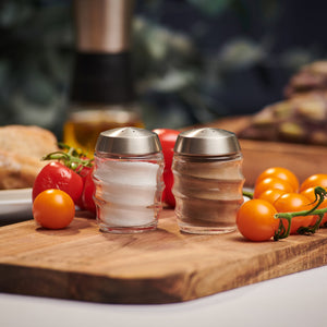 The Cole & Mason Bray Glass Shakers Set, featuring two glass shakers with stainless steel lids filled with salt and pepper, sits on a wooden board surrounded by red and yellow cherry tomatoes in a cozy kitchen.
