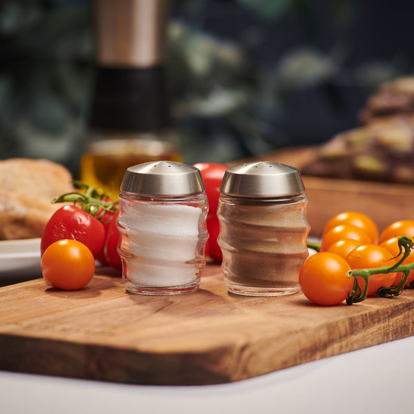 The Cole & Mason Bray Glass Shakers Set, featuring two glass shakers with stainless steel lids filled with salt and pepper, sits on a wooden board surrounded by red and yellow cherry tomatoes in a cozy kitchen.