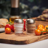 The Cole & Mason Bray Glass Shakers Set, featuring two glass shakers with stainless steel lids filled with salt and pepper, sits on a wooden board surrounded by red and yellow cherry tomatoes in a cozy kitchen.