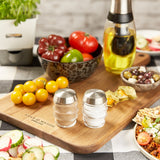 The Cole & Mason Bray Glass Shakers Set sits on a wooden cutting board alongside fresh tomatoes, an oil dispenser, olives, and mixed vegetables on a checkered tablecloth in a cozy kitchen.