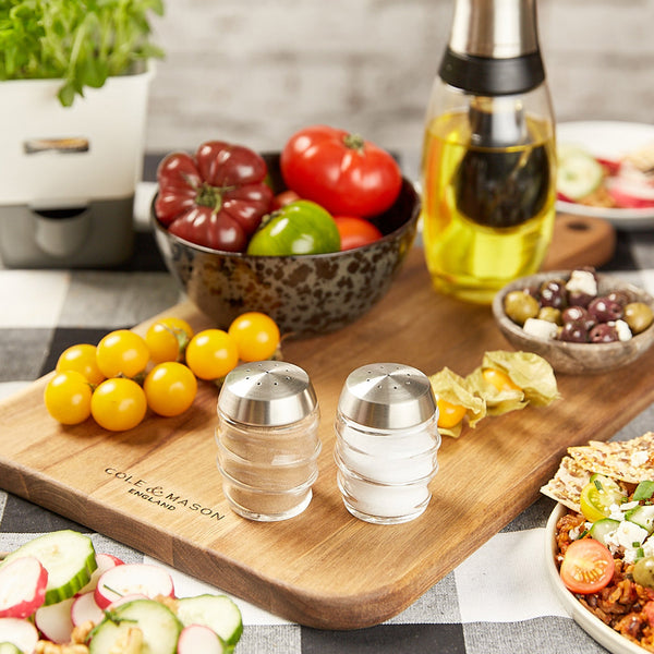 The Cole & Mason Bray Glass Shakers Set sits on a wooden cutting board alongside fresh tomatoes, an oil dispenser, olives, and mixed vegetables on a checkered tablecloth in a cozy kitchen.