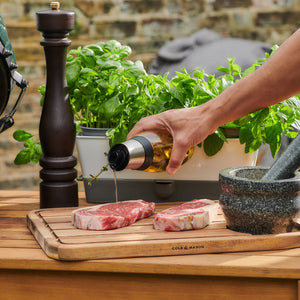 A hand uses the Cole & Mason Bristol Duo Oil & Vinegar Pourer for mess-free pouring onto two raw steaks on a wooden cutting board, with a pepper grinder, mortar and pestle, and fresh basil in a pot nearby on an outdoor wooden table.