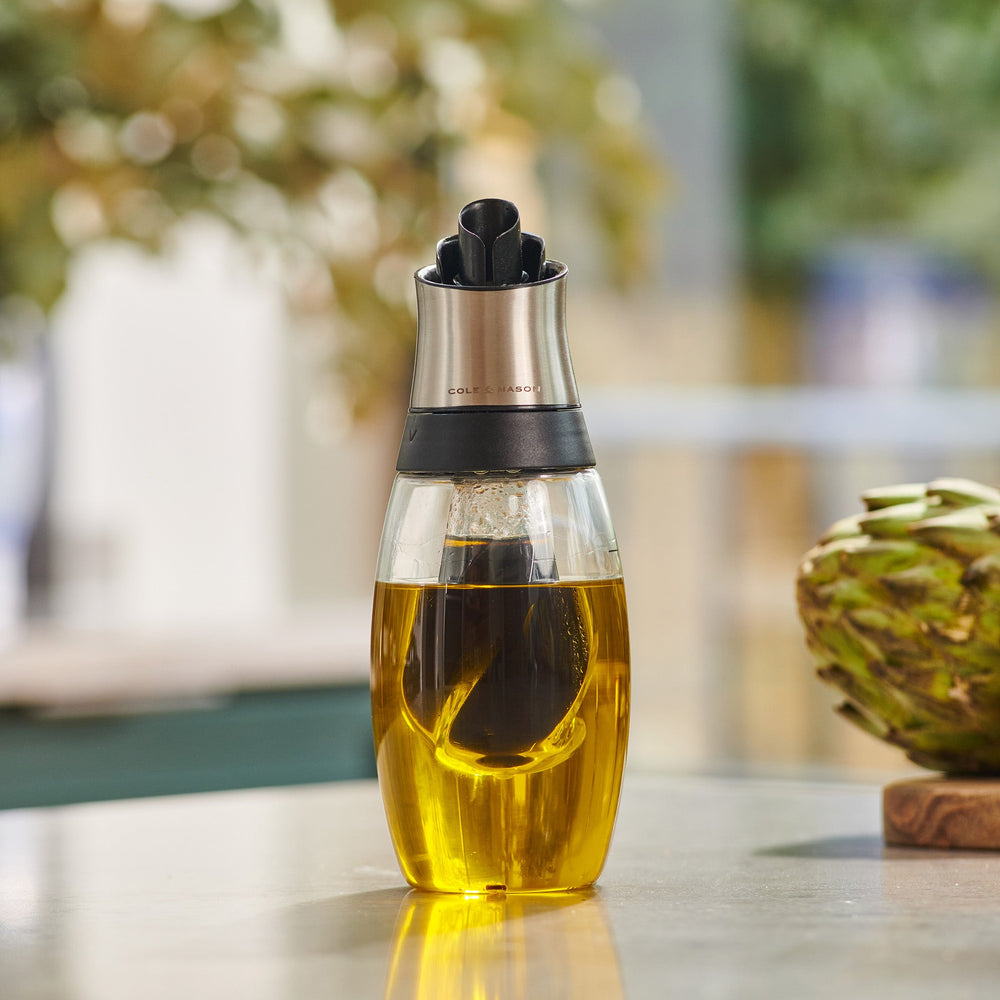 The Cole & Mason Bristol Duo Oil & Vinegar Pourer features clear glass with a metal top and black spout for mess-free dispensing. Filled with golden oil and a red chili pepper, it casts a soft shadow on a light background.