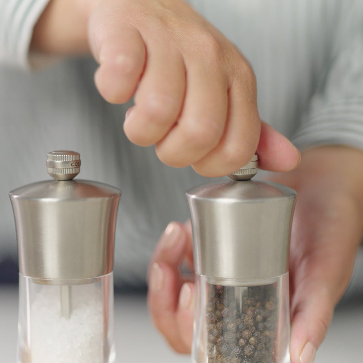 A person’s hands hold and twist the tops of two grinders, one containing coarse salt and the other black peppercorns, over a white surface.