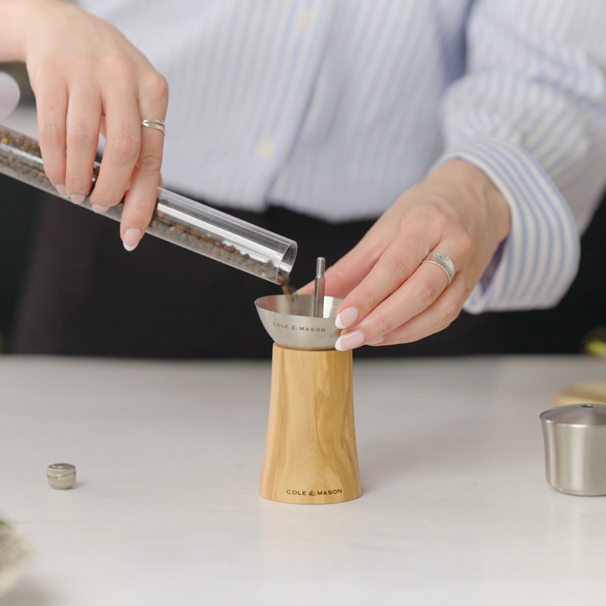 A person pours peppercorns from a glass tube into the top of a wooden Cole & Mason pepper grinder on a white surface.