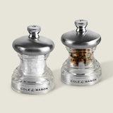 The Cole & Mason Button Salt & Pepper Mill Set features two clear grinders with metal tops labeled S and P. Compact and adjustable, one is filled with salt and the other with peppercorns, shown on a pale background.