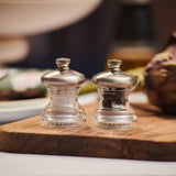 The Cole & Mason Button Salt & Pepper Mill Set, with clear bodies and silver tops plus adjustable coarseness, rests on a wooden board; blurred food and tableware are visible in the background.