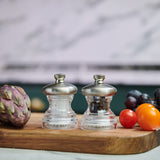 The Cole & Mason Button Salt & Pepper Mill Set features a compact glass design on a wooden cutting board with an artichoke, grapes, and cherry tomatoes. Adjustable coarseness settings make seasoning easy and precise.