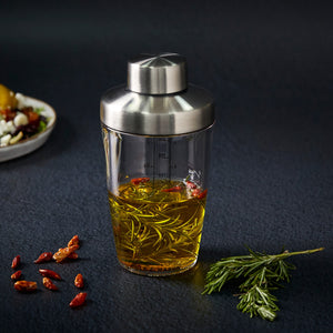 The Cole & Mason Cambourne Salad Dressing Shaker, featuring a glass body and metal lid, is displayed with olive oil, rosemary, and chili peppers—ideal for homemade dressings. A blurred plate of food sits in the background.
