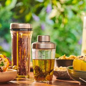 The Cole & Mason Cambourne Salad Dressing Shaker, filled with oil and herbs, sits by a salad shaker and fresh food bowls on a table—ideal for making vinaigrettes—with a blurred green garden in the background.