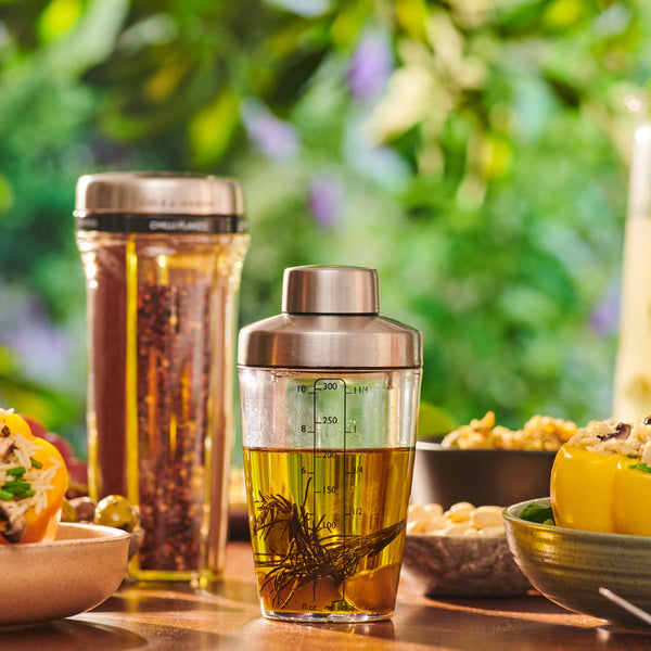 The Cole & Mason Cambourne Salad Dressing Shaker, filled with oil and herbs, sits by a salad shaker and fresh food bowls on a table—ideal for making vinaigrettes—with a blurred green garden in the background.