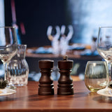 The Cole & Mason London Chocolate Wood Salt Shaker & Pepper Mill Set, crafted from natural beech wood, sits on a wooden table with empty glassware nearby and a blurred dining scene in the background.