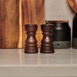 Two Cole & Mason London Chocolate Wood Salt Shaker & Pepper Mill sit side by side on a kitchen countertop, with a cutting board and other kitchen items in the background.