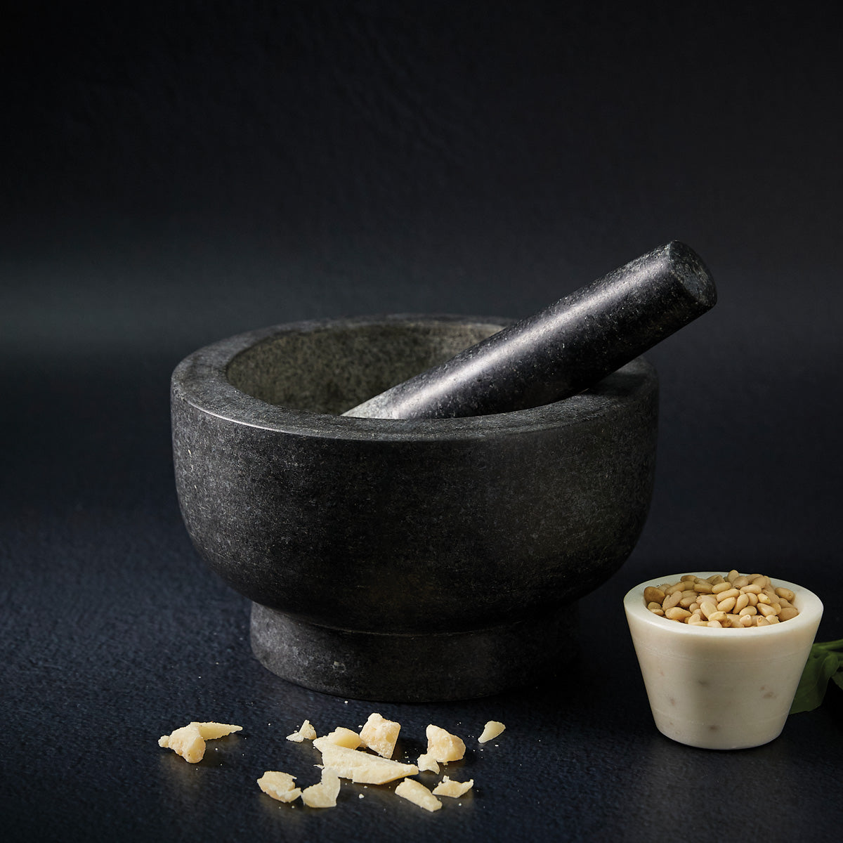 A dark stone mortar and pestle sit on a black surface, with some crushed pieces nearby. Next to them is a small white bowl filled with seeds or grains.