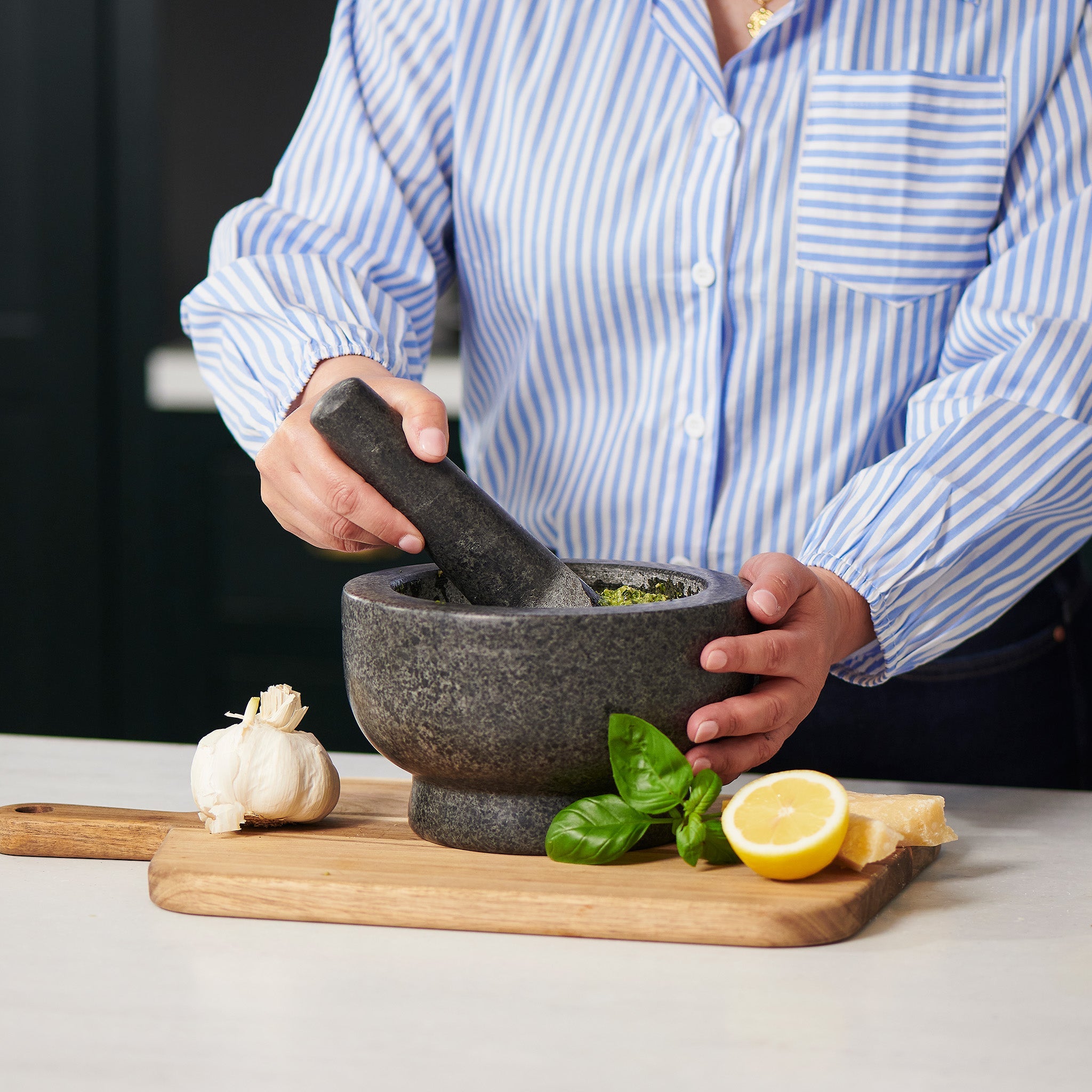 A home chef in a blue striped shirt uses the Cole & Mason Clavering Pestle & Mortar on a wooden board with garlic, basil, lemon, and cheese nearby to maximize grinding efficiency.