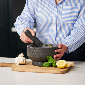 A home chef in a blue striped shirt uses the Cole & Mason Clavering Pestle & Mortar on a wooden board with garlic, basil, lemon, and cheese nearby to maximize grinding efficiency.