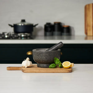 The Cole & Mason Clavering Pestle & Mortar rests on a wooden board with garlic, basil, lemon, and cheese in a modern kitchen—ideal for home chefs who value superior grinding efficiency.