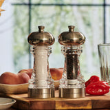 The Cole & Mason Clovelly Salt & Pepper Mill Set, with a modern clear design and adjustable grinding mechanisms, sits on a wooden countertop among peaches, red peppers, and a glass, framed by a blurred window.