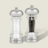 The Cole & Mason Clovelly Salt & Pepper Mill Set features two transparent mills with metallic tops—one containing white salt crystals, the other black peppercorns—both showcasing a modern design and adjustable grinding mechanisms on a light background.