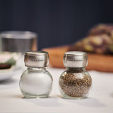 Two round glass shakers with stainless steel lids, labeled Cole & Mason, sit together. The Darlington Shakers Set combines classic glass and stainless steel for stylish seasoning at any meal.