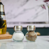 Two glass spice jars with stainless steel lids from the Cole & Mason Darlington Shakers Set, one with salt and the other with ground black pepper, sit side by side on a marble countertop, displaying their elegant design.