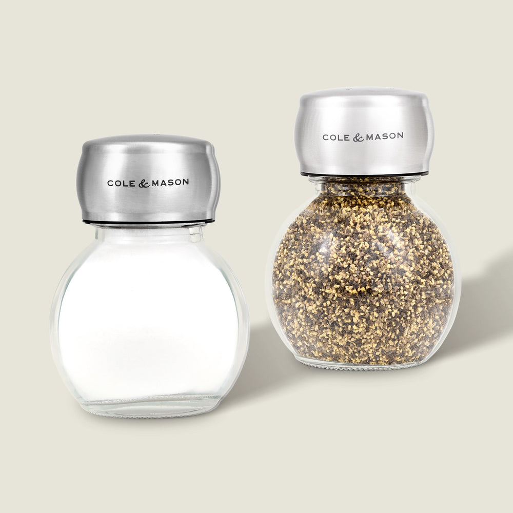 The Cole & Mason Darlington Shakers Set includes two glass jars with stainless steel lids—one empty, one filled with cracked black pepper. Both feature a sleek design and Cole & Mason branding, perfect for stylish seasoning.