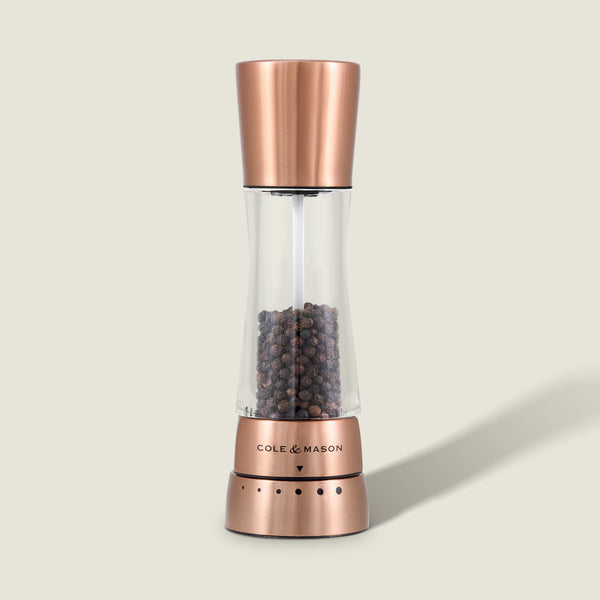 A Cole & Mason Derwent Copper Salt & Pepper Mill, filled with black peppercorns and featuring precision grind technology, stands upright on a light beige background.