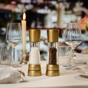 Cole & Mason Derwent Gold Salt & Pepper Mills, with gold-tone stainless steel tops and advanced grinding technology, grace a formal dining table set with crystal glasses, candlelight, and white napkins in a refined atmosphere.