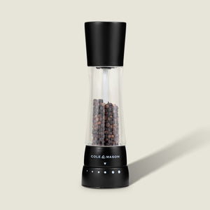 #pepper A Cole & Mason Derwent Matt Black Salt & Pepper Mill with an acrylic body and adjustable grind settings, filled with whole peppercorns, stands upright on a light beige surface with a subtle shadow to the right.