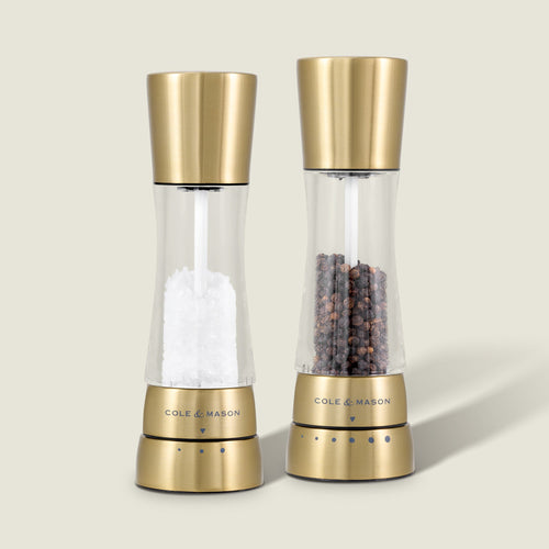 The Cole & Mason Derwent Gold Salt & Pepper Mills, filled with white salt crystals and black peppercorns, stand side by side on a neutral background, highlighting their advanced grinding technology.