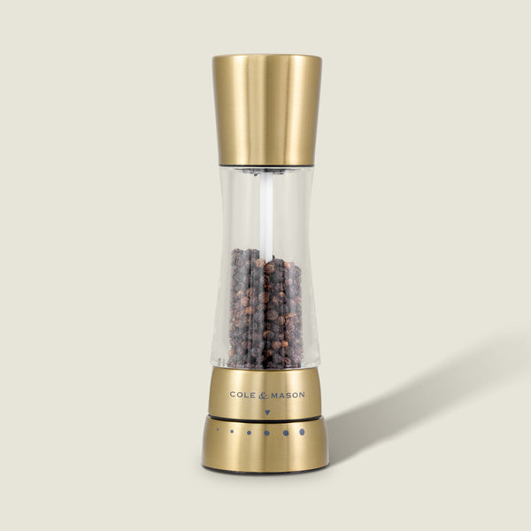 The Cole & Mason Derwent Gold Salt & Pepper Mill features gold-tone stainless steel, advanced grinding technology, and is filled with black peppercorns, all set against a plain, light background.