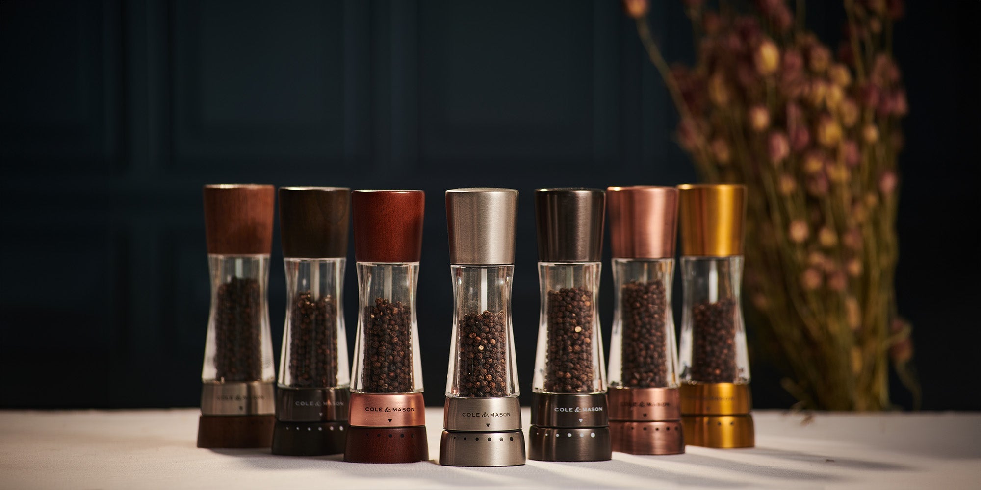 A row of six pepper grinders in various metallic and wood finishes sits on a white surface, with dried flowers blurred in the background. Each grinder is filled with whole peppercorns.