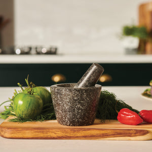 The Cole & Mason Dorchester Granite Pestle & Mortar sits on a wooden board with green tomatoes, fresh herbs, and two red chili peppers—ideal for grinding herbs and making flavorful spice blends in your kitchen.