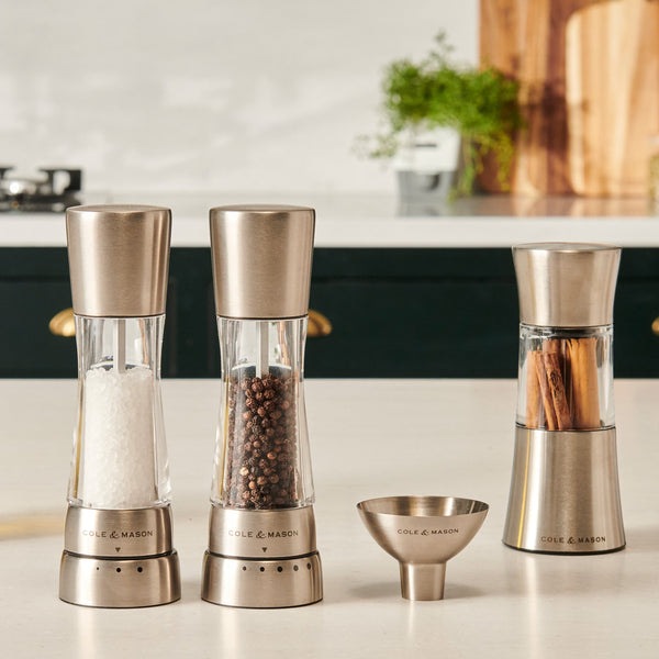Three Cole & Mason stainless steel spice grinders with salt, pepper, and cinnamon sticks sit beside the Dover Stainless Steel Funnel on the counter. Their sleek, easy-to-clean design gleams against the blurred kitchen backdrop.