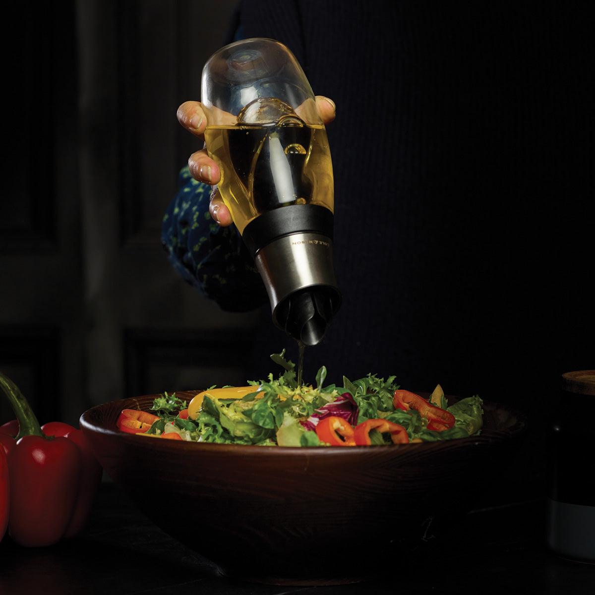 A person pours oil from a bottle with a spout over a fresh salad in a wooden bowl, with colorful bell peppers visible nearby.