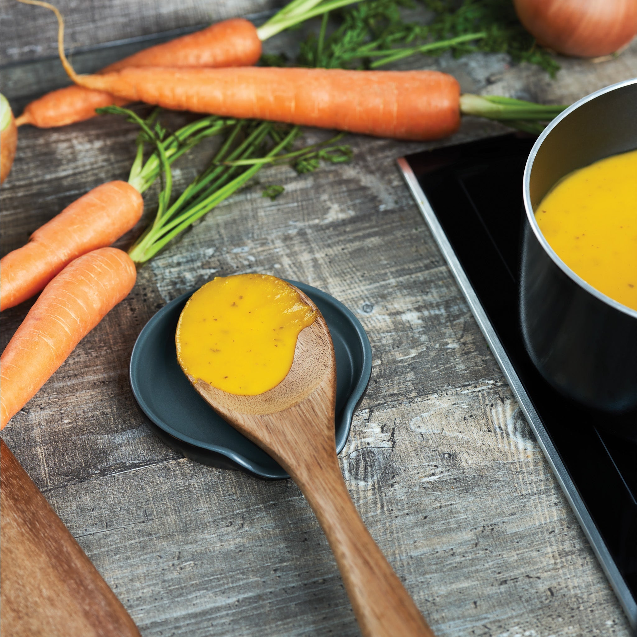 A wooden spoon with creamy orange soup rests on a small dish, next to fresh carrots and a pot of soup on a stovetop, all on a rustic wooden surface.