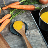 A wooden spoon with creamy orange soup rests on the Cole & Mason Duxford Ceramic Spoon Rest beside a pot of soup, surrounded by fresh carrots and an onion—a stylish, functional accent for your kitchen decor.
