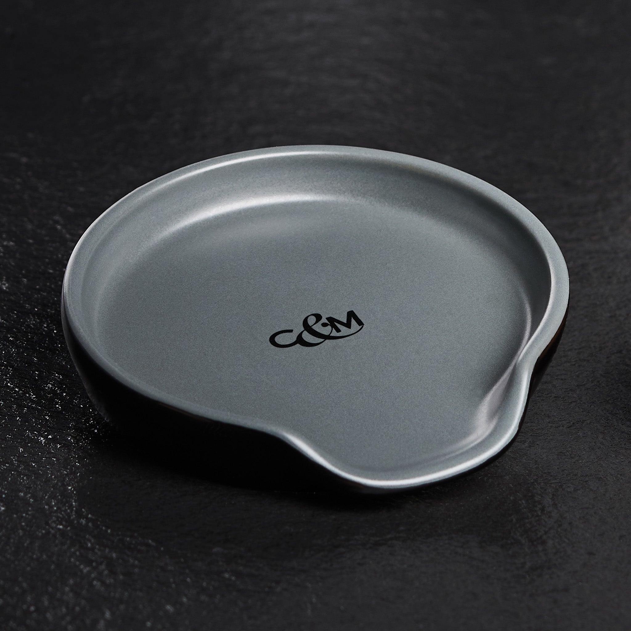 A round, matte gray dish with a small spout and the initials C&M printed in black in the center, placed on a textured dark surface.