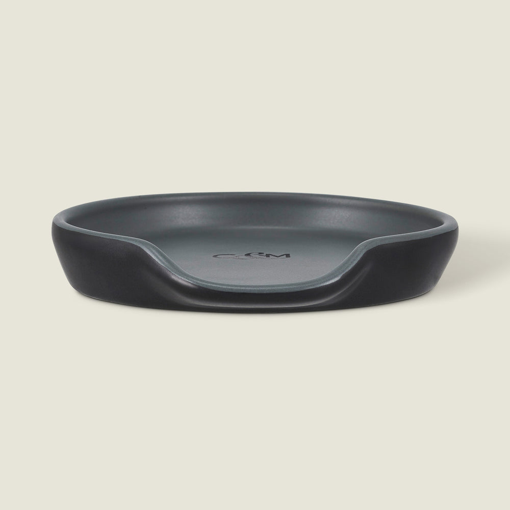 The Cole & Mason Duxford Ceramic Spoon Rest is a black, round ceramic piece with a raised edge and front cutout—perfect for kitchen decor and shown against a light cream background.