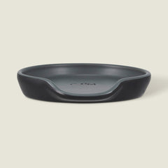The Cole & Mason Duxford Ceramic Spoon Rest is a black, round ceramic piece with a raised edge and front cutout—perfect for kitchen decor and shown against a light cream background.