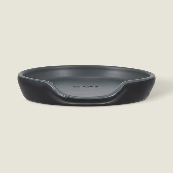 The Cole & Mason Duxford Ceramic Spoon Rest is a black, round ceramic piece with a raised edge and front cutout—perfect for kitchen decor and shown against a light cream background.
