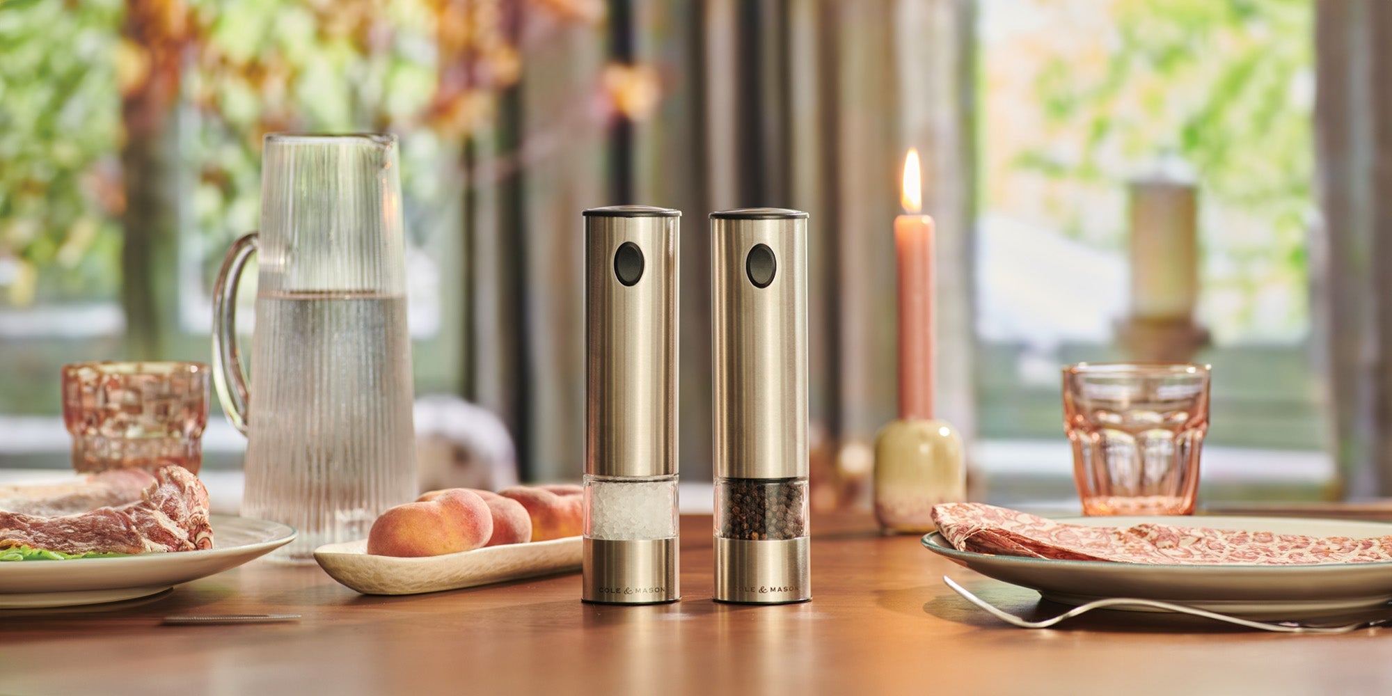 Two metallic salt and pepper grinders sit on a wooden dining table set with plates of food, a glass pitcher of water, a candle, glasses, and a dish of peaches. Sunlight filters through a window in the background.