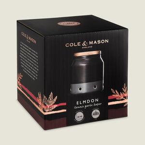 A black box with copper accents labeled Cole & Mason features the Elmdon Ceramic Garlic Keeper—a stylish ceramic jar with a wooden lid, perfect for storing garlic and adding elegance to any kitchen.