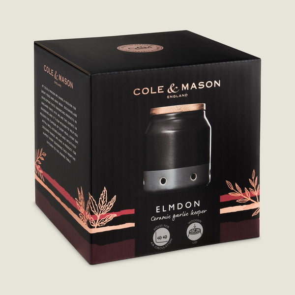 A black box with copper accents labeled Cole & Mason features the Elmdon Ceramic Garlic Keeper—a stylish ceramic jar with a wooden lid, perfect for storing garlic and adding elegance to any kitchen.