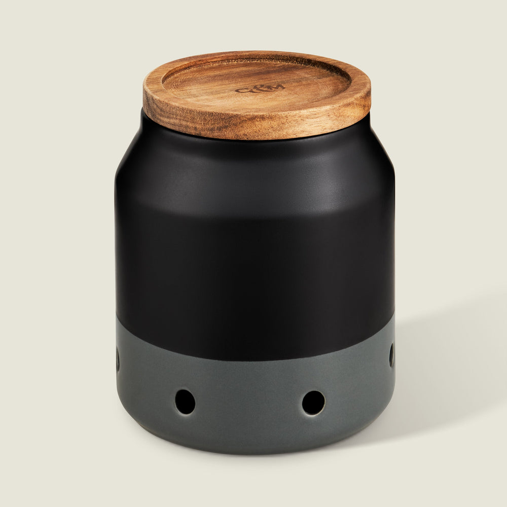 The Cole & Mason Elmdon Ceramic Garlic Keeper, in black and gray with a wooden lid and round ventilation holes, offers stylish garlic storage and appears against a light background.