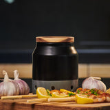 The Cole & Mason Elmdon Ceramic Garlic Keeper, featuring a black and silver design with a wooden lid, sits on a wooden surface beside garlic bulbs, lemon wedges, and grilled skewers—an elegant kitchen accent against a dark background.